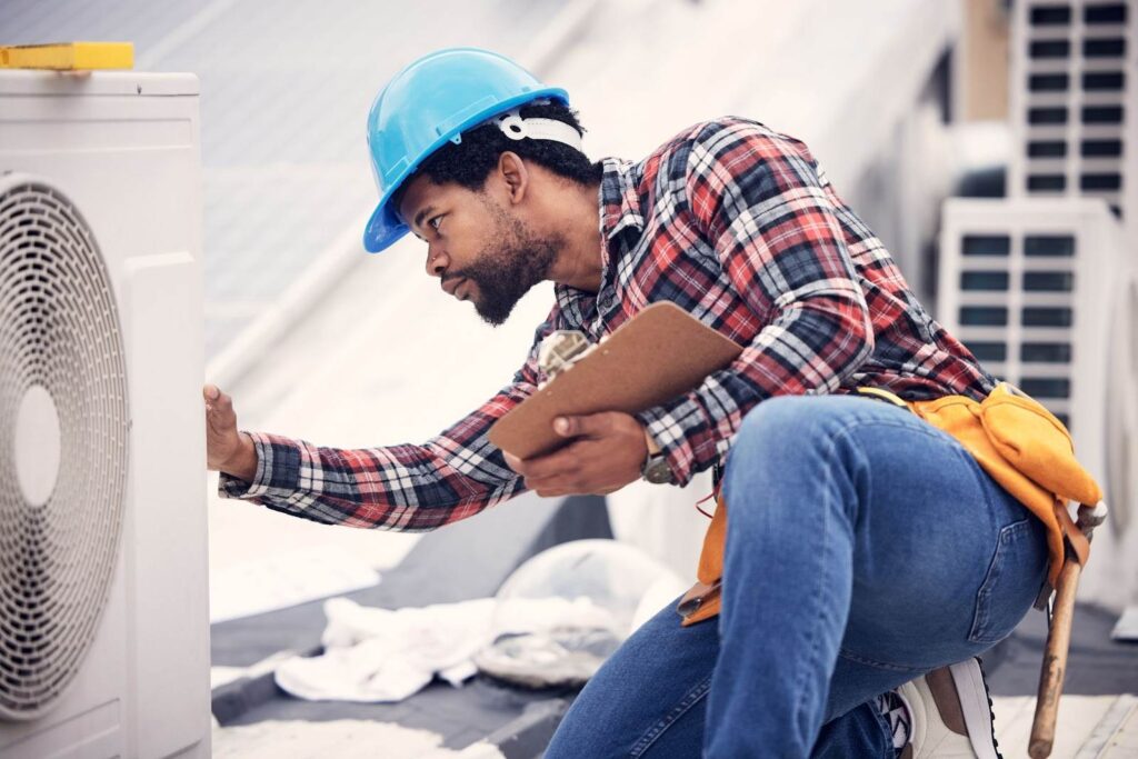 A worker in a blue hard hat and plaid shirt inspects an air conditioning unit on a rooftop, holding a clipboard and wearing a tool belt.